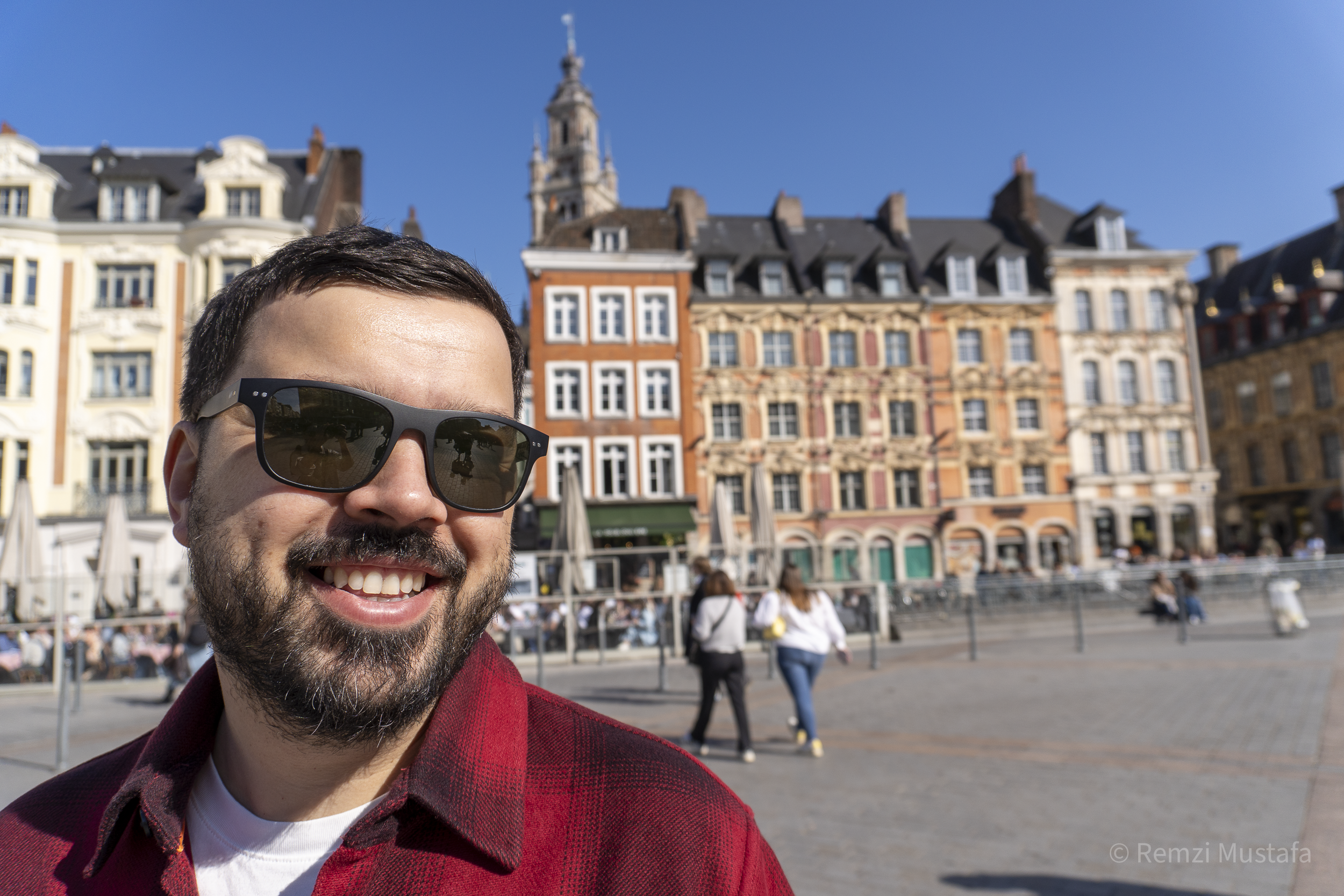 A smiling man with dark hair, beard, and sunglasses taking a selfie in a European city square with colorful buildings and pedestrians in the background.