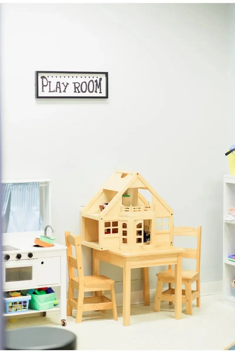 A children’s playroom with a miniature wooden dollhouse on a small table, surrounded by three small wooden chairs, with a sign reading 'Play Room' on the wall.