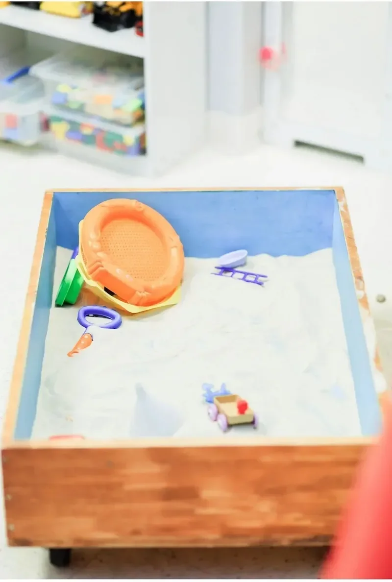 Silhouette of a sandbox with sand toys including a yellow bucket, purple shovel, green scoop, and a small wooden toy cart, set indoors.