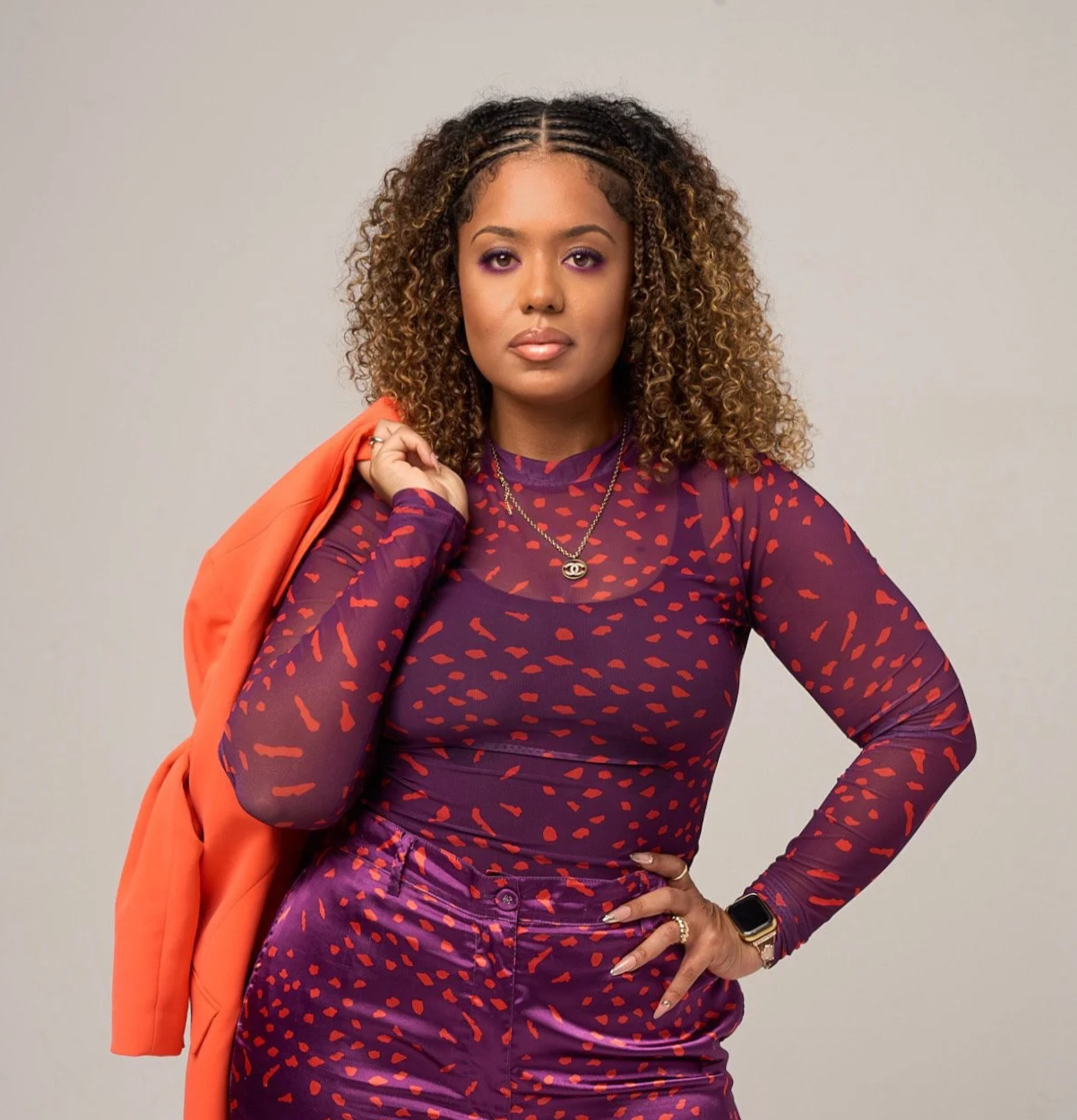 A woman with curly hair and purple eyeshadow wearing a purple and red patterned top and matching pants, holding an orange jacket over her shoulder, and posing against a plain light-colored background.