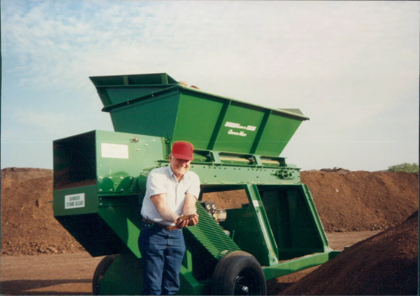A man in a white shirt, blue pants, red hat, and gloves standing in front of a large green industrial machine outdoors on a dirt field, with piles of dirt behind him and a partly cloudy sky.