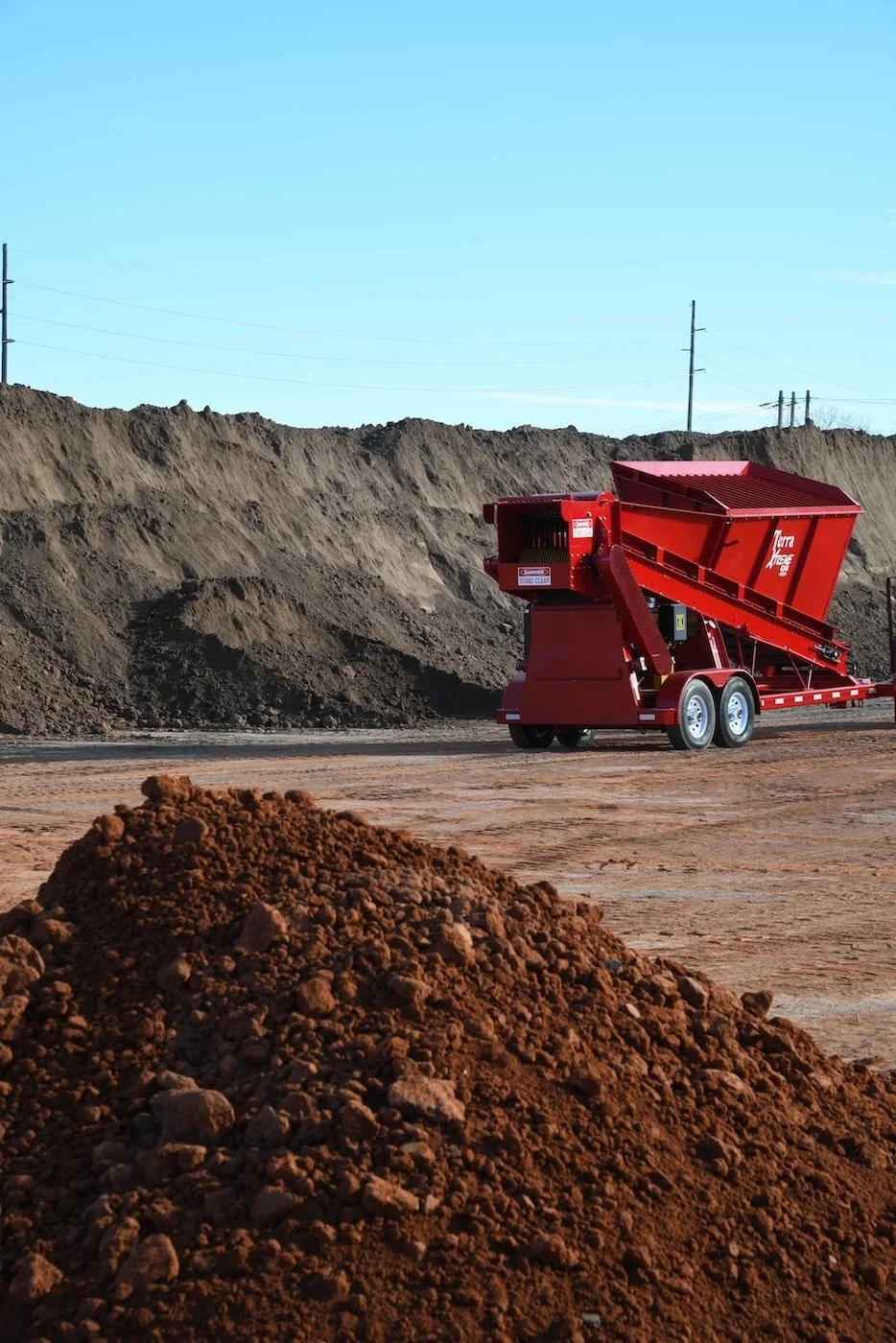 A large red soil splitter on wheels at a construction site with piles of dirt and soil in the background under a blue sky.