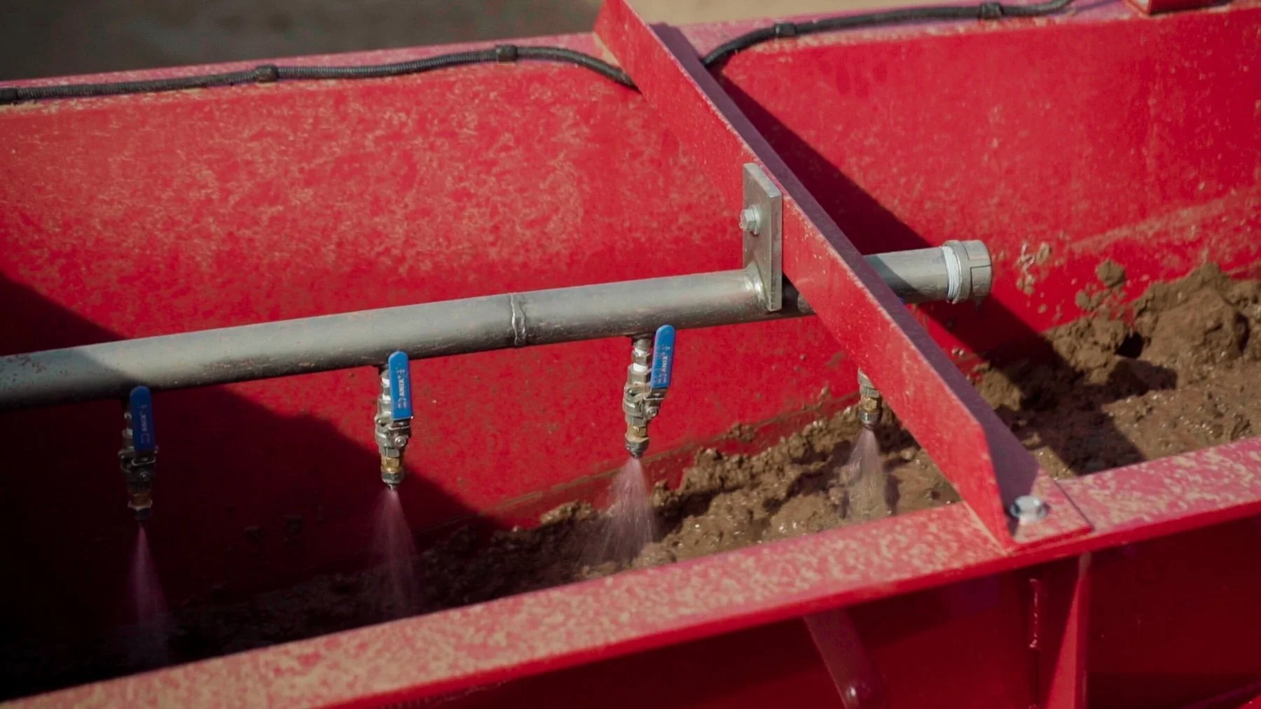 Close-up of a red farming irrigation system with three blue valve handles connected to pipes, watering freshly tilled soil.