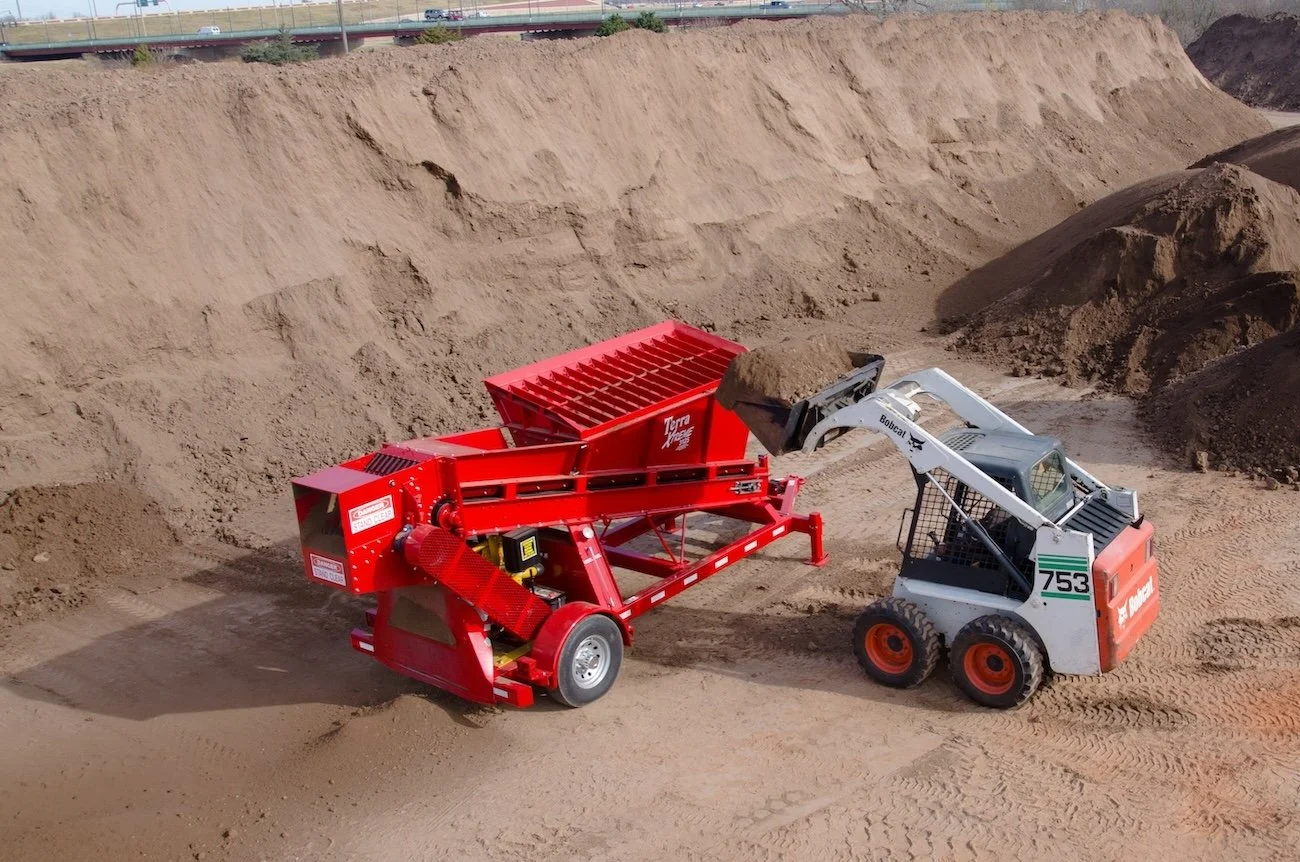 A construction site with a Bobcat skid-steer loader dumping dirt into a red Terra Track attachment, shaping a dirt embankment.