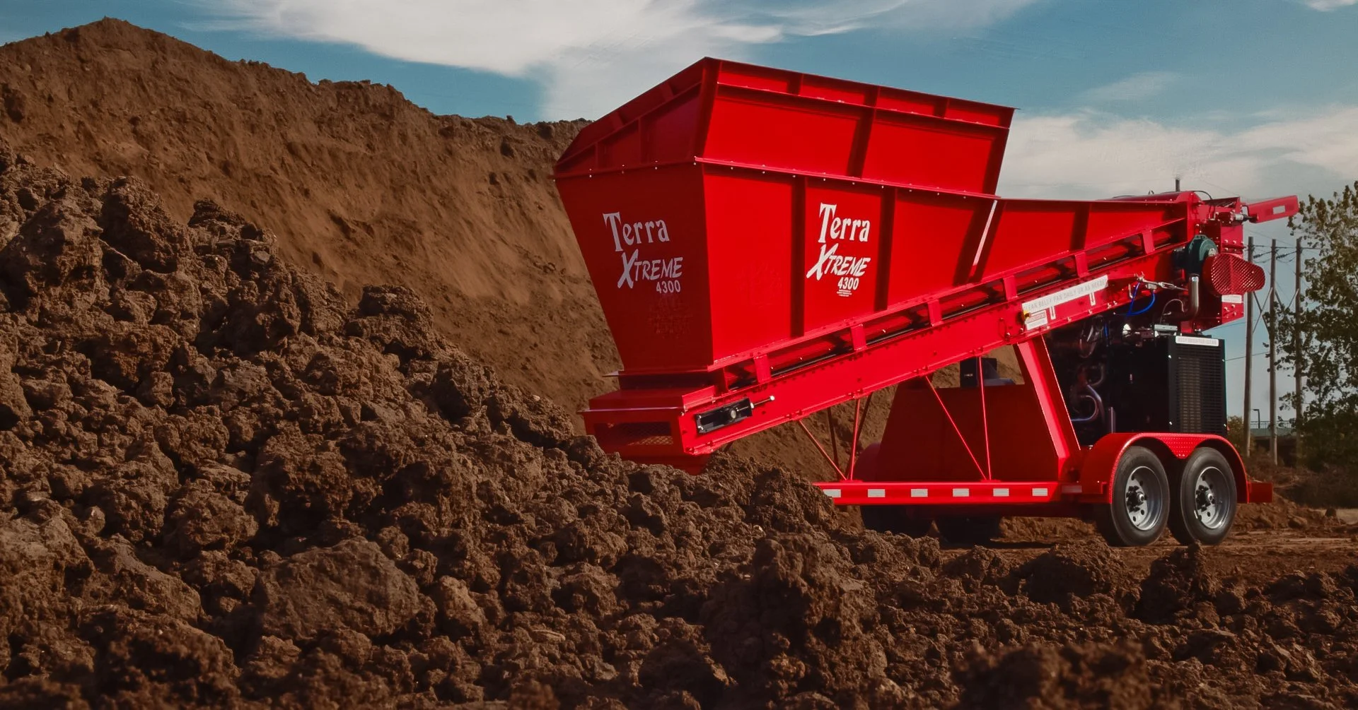 A red Terra Xtreme 4300 soil screener on a trailer, positioned next to a large pile of soil or dirt outdoors on a clear day.