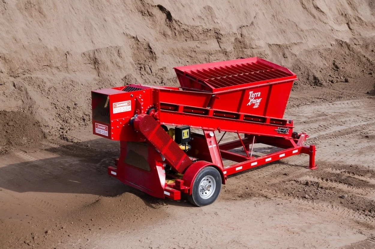 Red soil screener machine with large hopper and conveyor in a dirt excavation site.