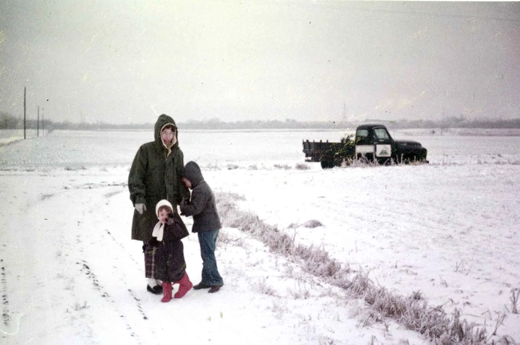 A person and two children standing outdoors in a snow-covered field, with an old black pickup truck in the background on the right.