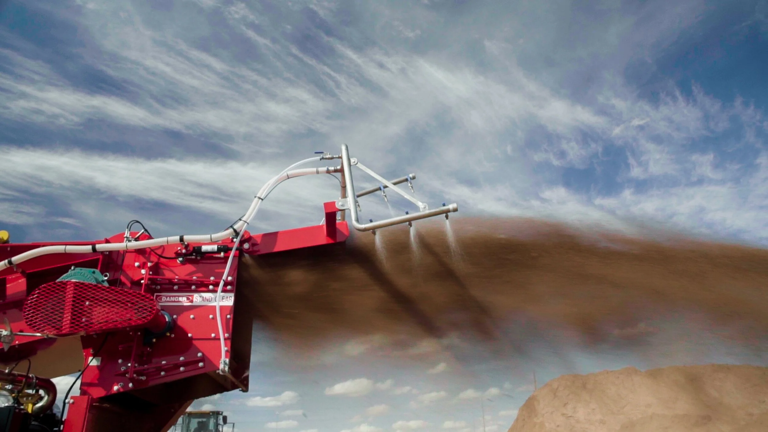 A large piece of red agricultural machinery spreading soil or compost in the air at a farm field with a partly cloudy sky in the background.