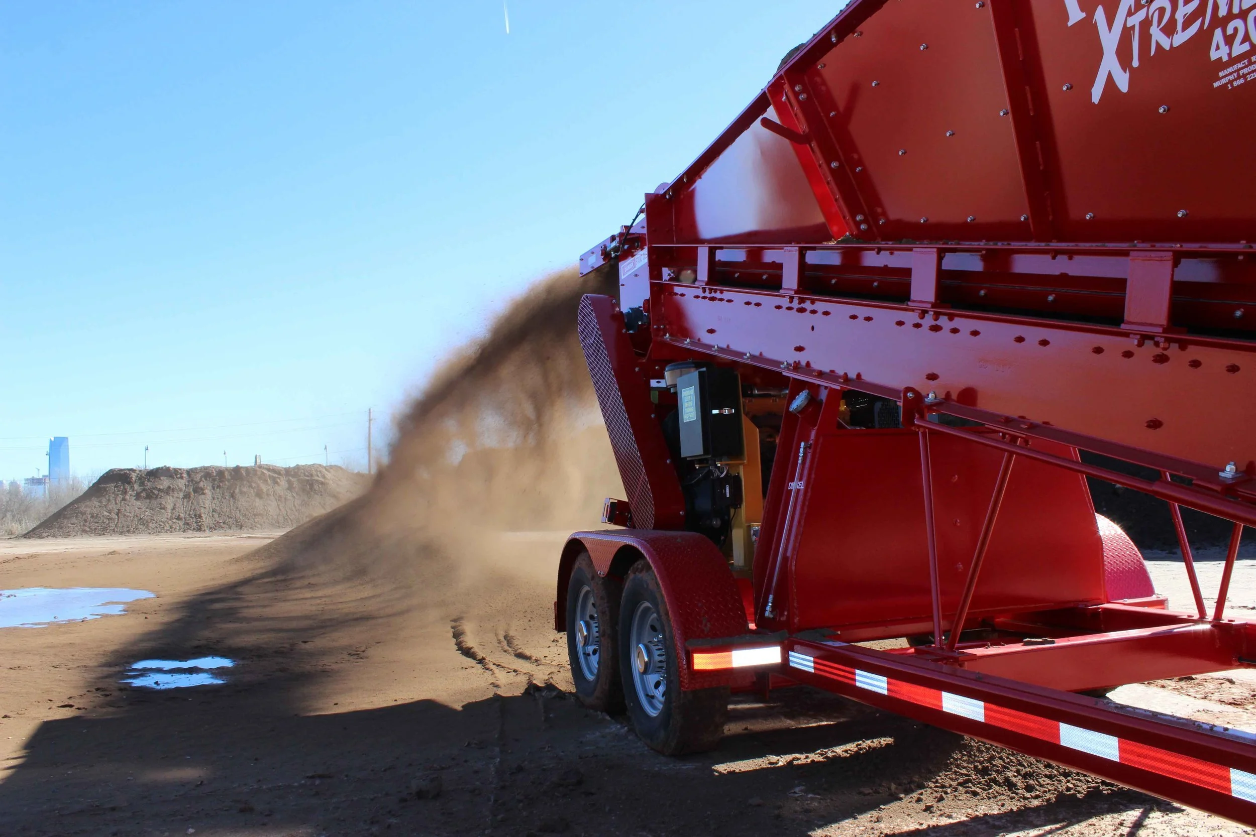 A large red industrial machine spreading dirt and soil on an outdoor construction or landscaping site under a clear blue sky.