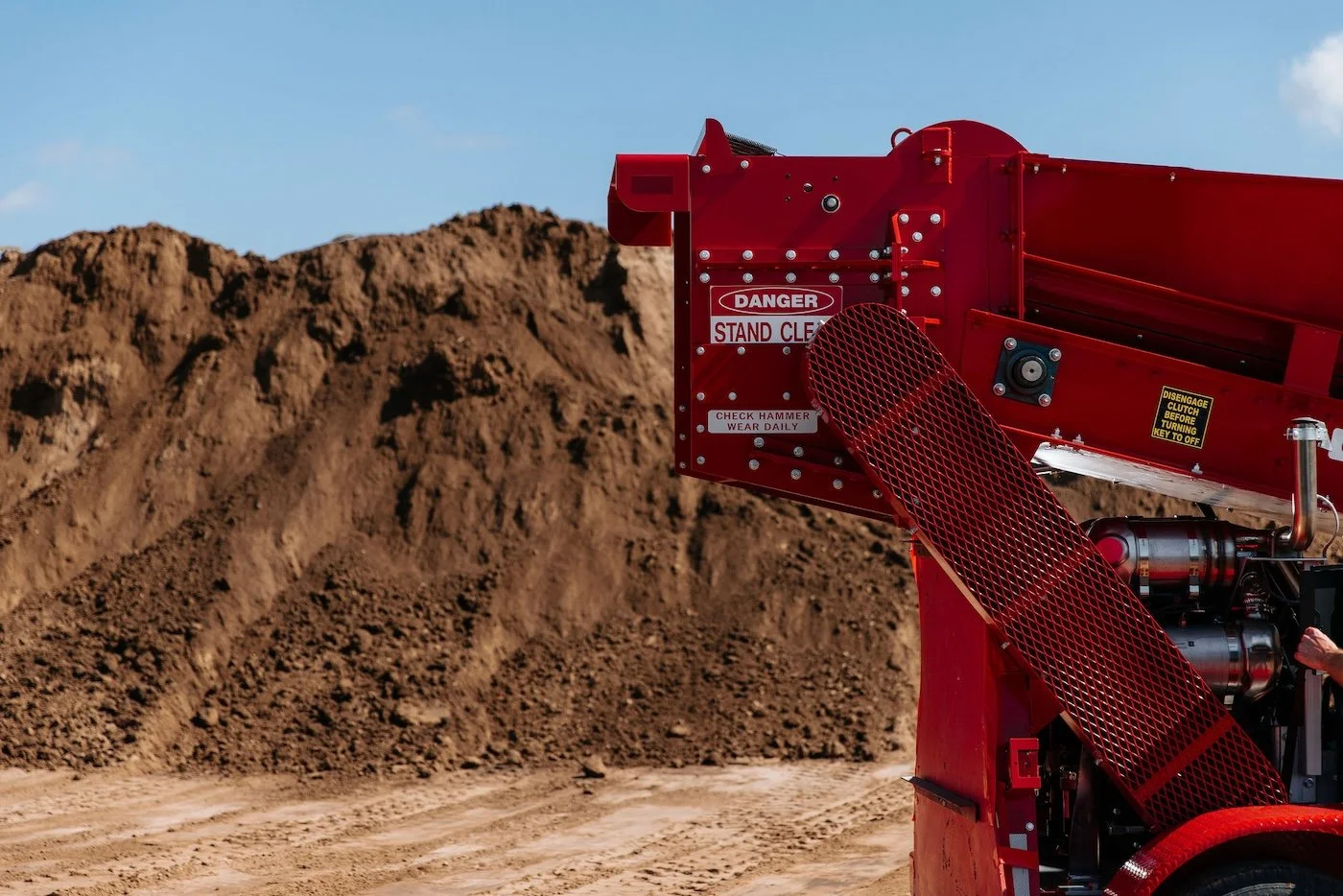 Close-up of a red industrial machine with warning signs, set against a background of dirt mounds and blue sky.