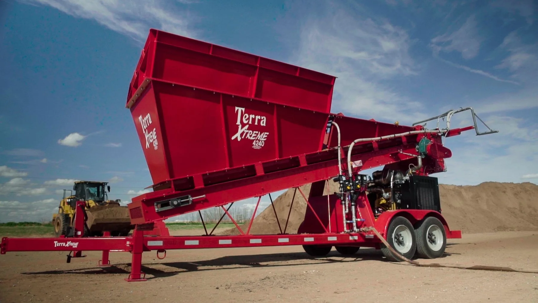 Red terraXtreme seed cleaner machine on a trailer with a large hopper and piping, set on a dirt field with a yellow bulldozer in the background, under partly cloudy sky.