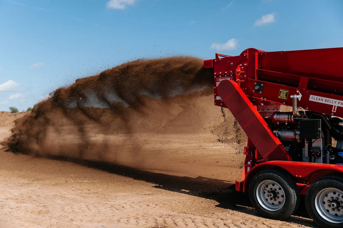 A red soil-moving machine dumping dirt on a sunny day with blue sky and clouds.
