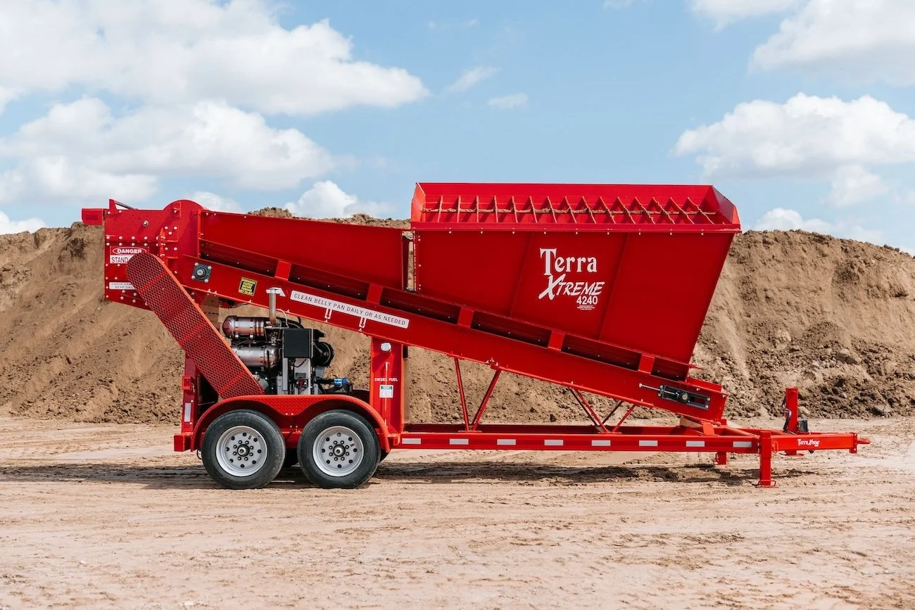 A red Terra XTREME 4240 soil screener on a dirt surface, with a large pile of dirt in the background and a partly cloudy sky.
