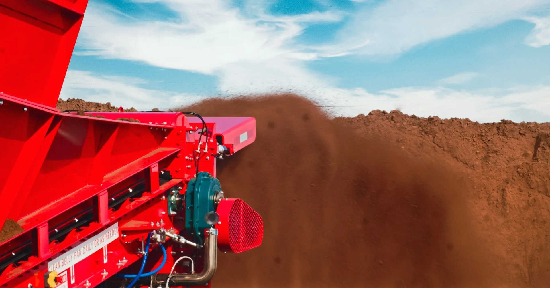 Close-up of a red soil auger machine displacing earth and dirt during land clearing, with a blue sky and clouds in the background.