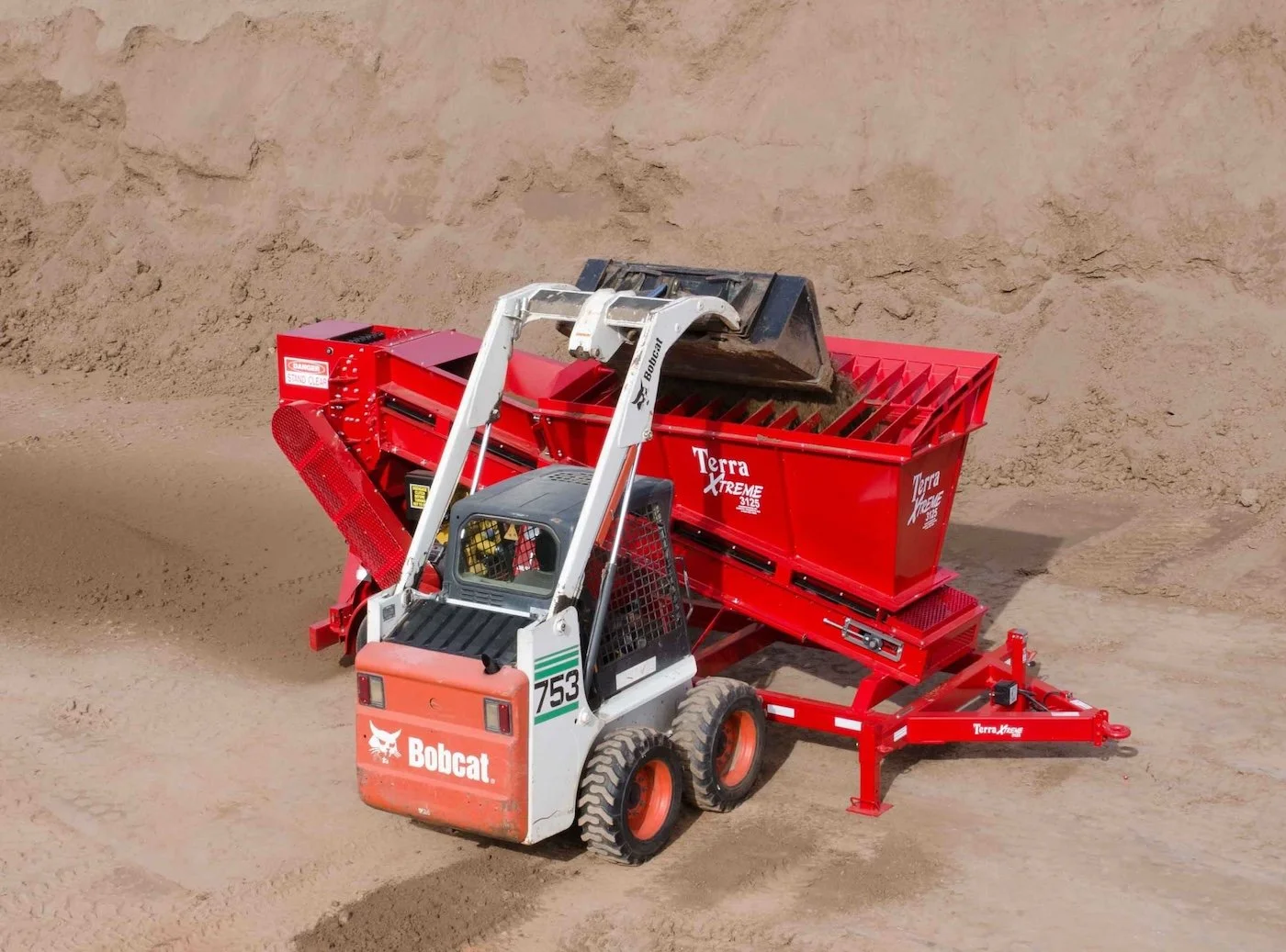 A Bobcat skid-steer loader with a Terra Xtreme soil screener attachment, working on a sandy construction site.