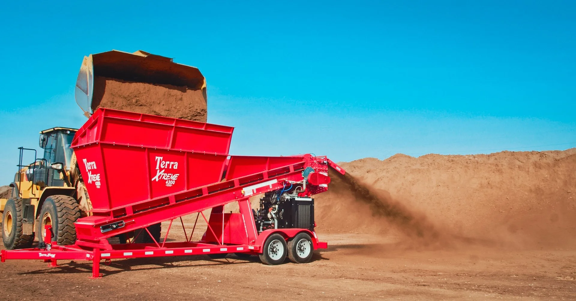 A red portable soil crusher pouring soil onto a large pile of dirt while a yellow loader loads soil into the crusher, set against a clear blue sky.