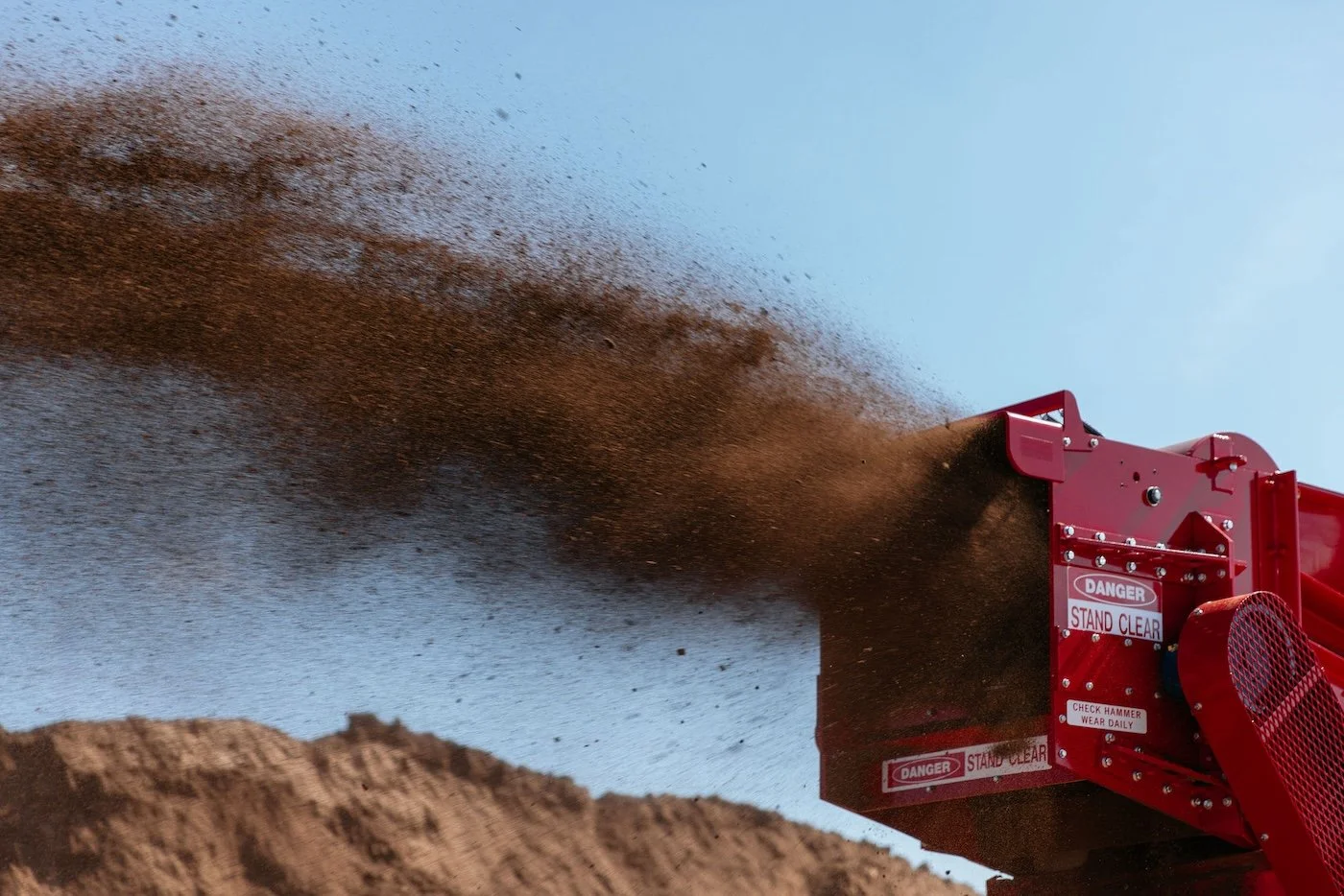 Close-up of an industrial red machine dispersing brown dirt or sand into the air against a blue sky.