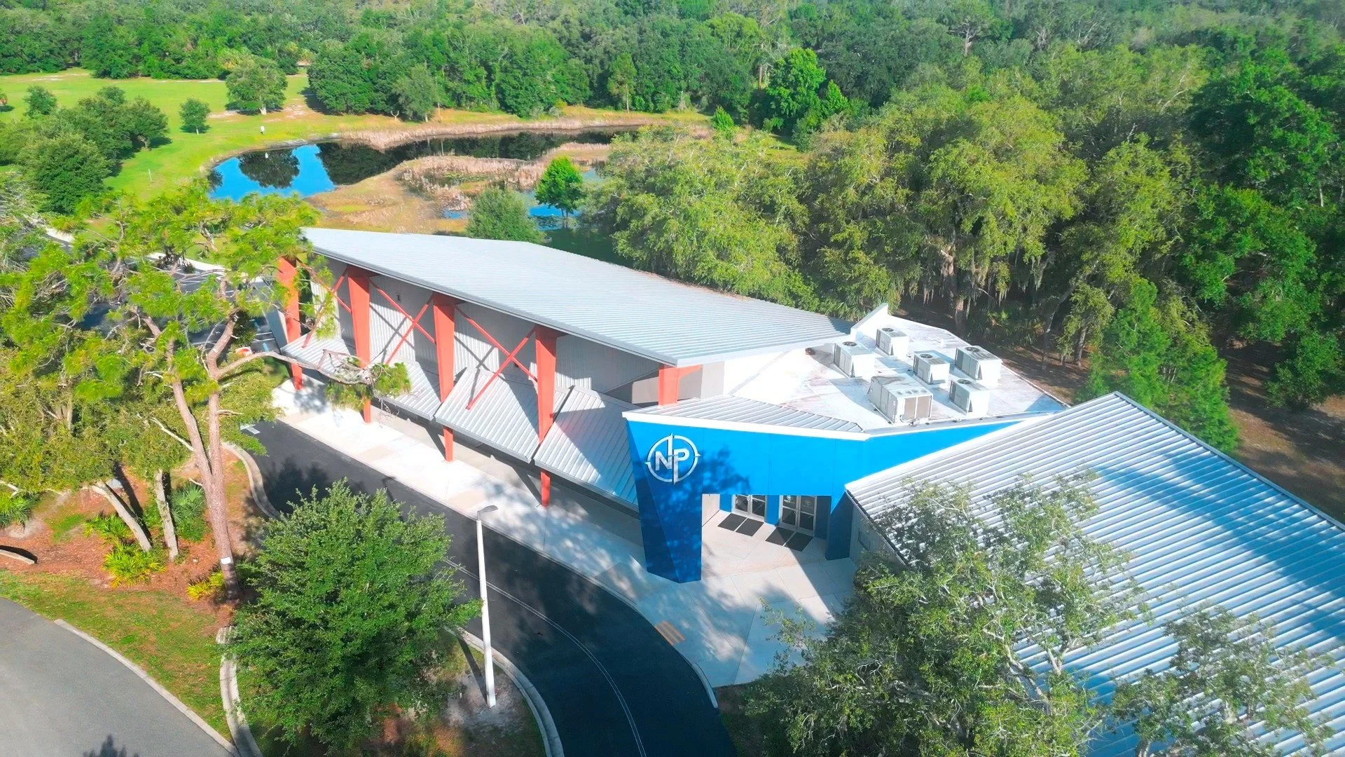 Aerial view of a modern building with a blue facade and a large parking lot, surrounded by lush green trees and a pond in the background.