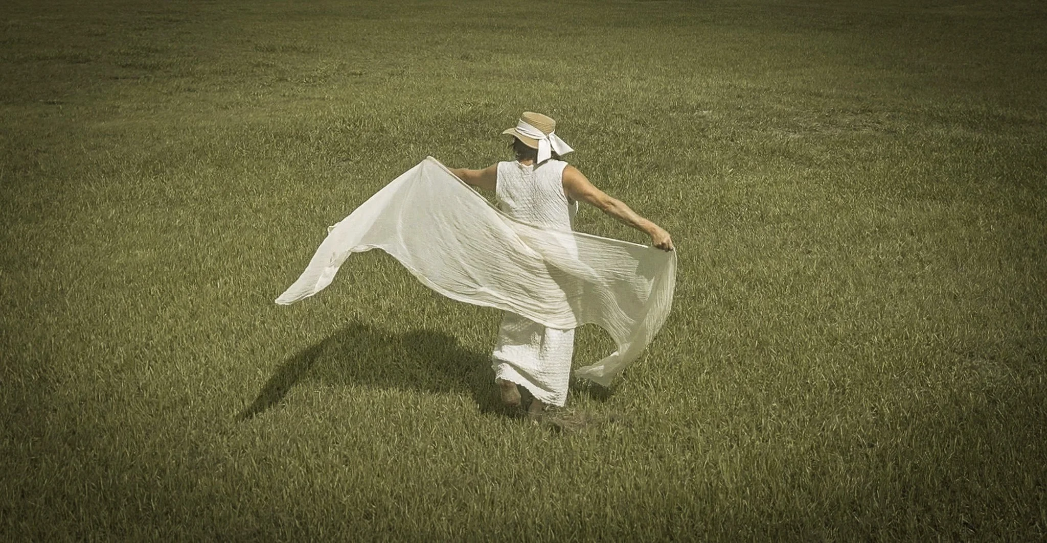 A woman in a white dress and hat holding a white cloth walking across a grassy field.
