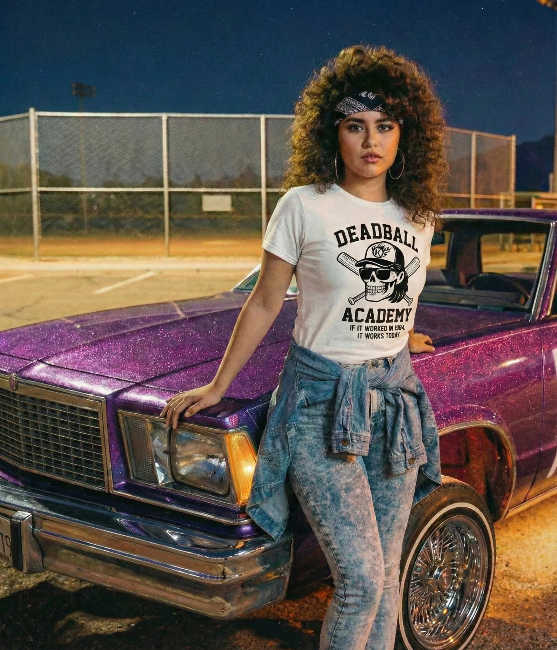 A woman with curly hair and a bandana leaning on a vintage purple car at a tennis court during dusk, wearing a white t-shirt with the words 'Deadball Academy' and a skull graphic, paired with acid-washed jeans and a denim jacket tied around her waist.