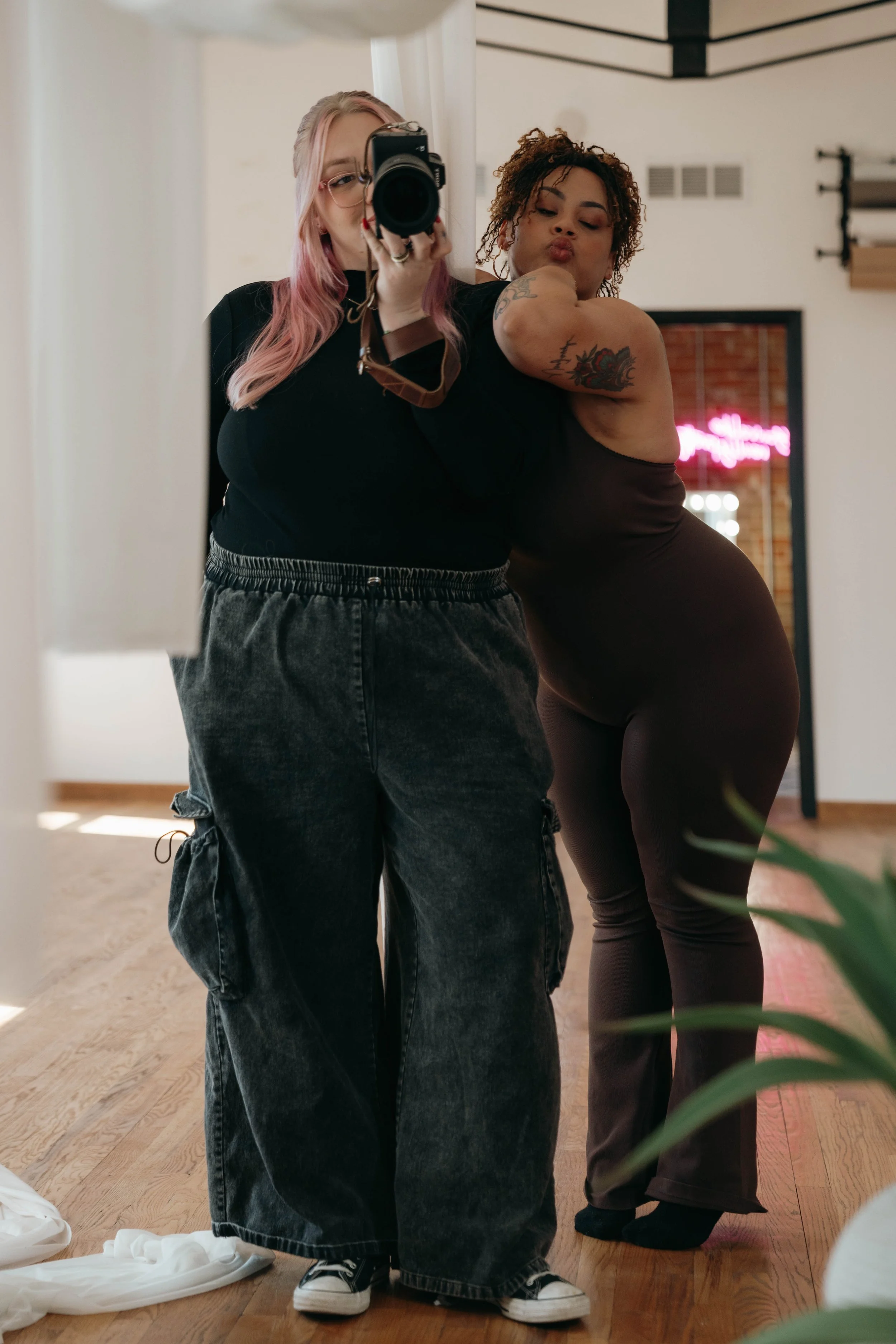 Two women taking a mirror selfie in a room with hardwood floors. The woman on the left has pink hair, glasses, and is wearing a black top and loose black jeans. The woman on the right has curly hair, tattoos, and is wearing a dark sleeveless body suit. She is making a kissy face and leaning toward the woman on the left.
