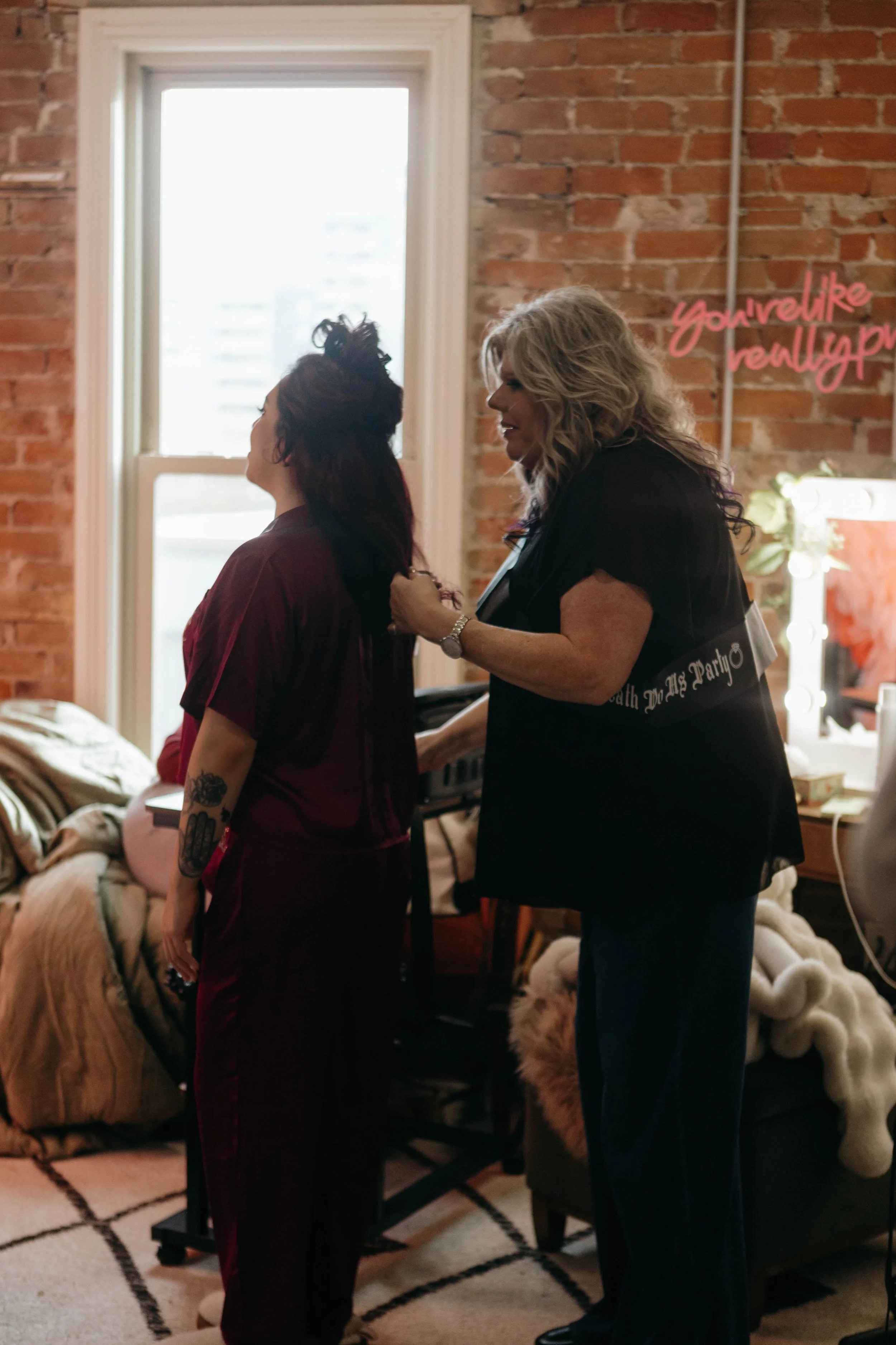 A woman with long gray hair helping another woman with long red hair prepare in a bedroom with an exposed brick wall. The woman with gray hair is wearing a black top and jeans, while the other woman is wearing maroon pajamas with tattoos on her arm. The room contains a bed with blankets, a window letting in natural light, and a neon sign on the wall that reads 'You look like really P...'