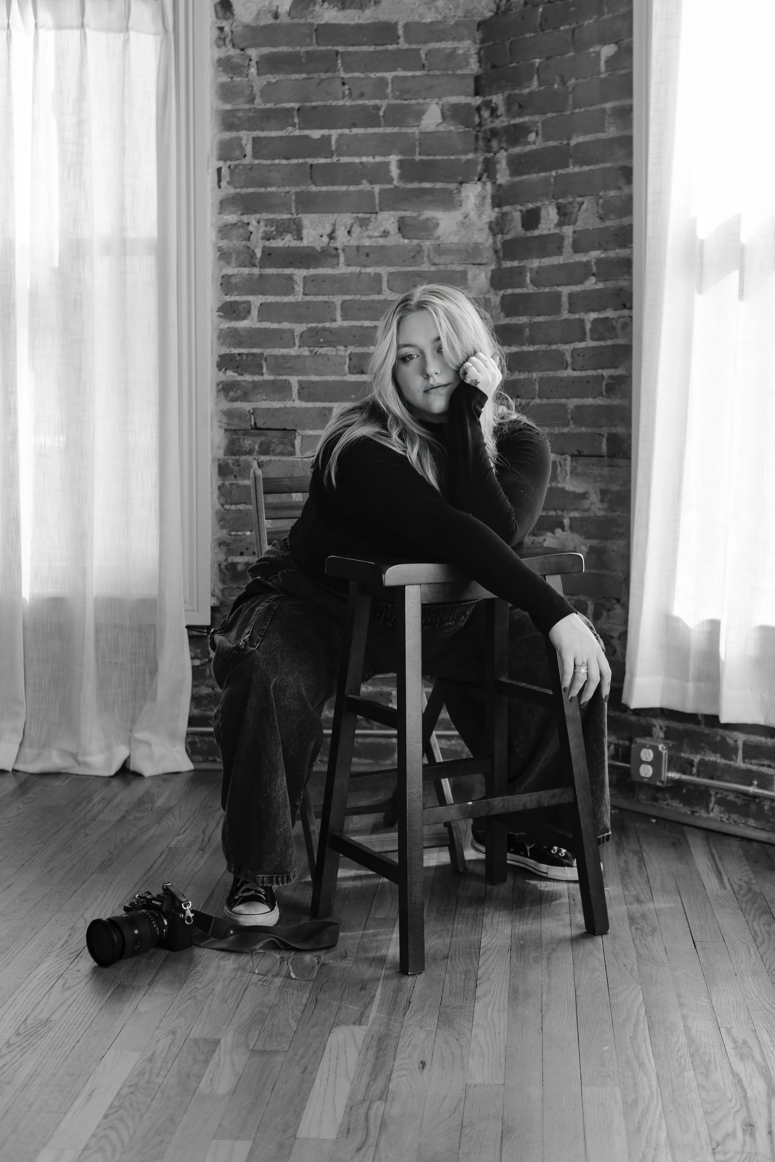 A woman sitting on a chair with her chin resting on her hand, looking at the camera in a room with exposed brick wall and large windows with curtains, with a camera on the floor nearby.