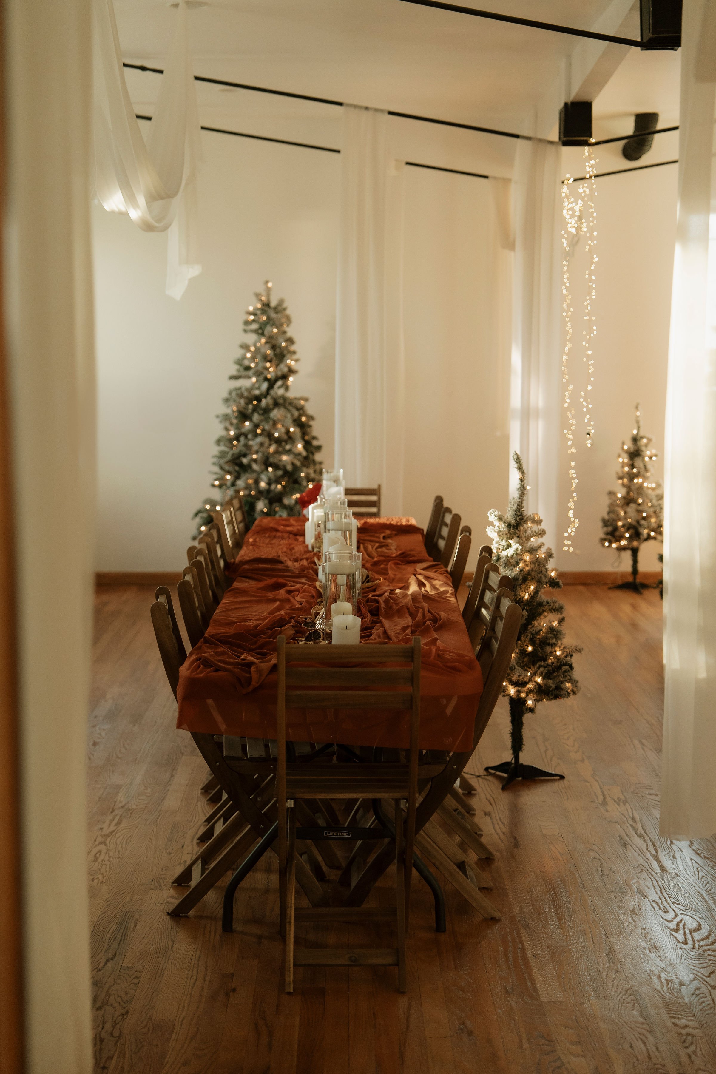 A long wooden dining table covered with an orange tablecloth, set with candles and decorative items, surrounded by wooden chairs, with Christmas trees and fairy lights in the background in a decorated room.