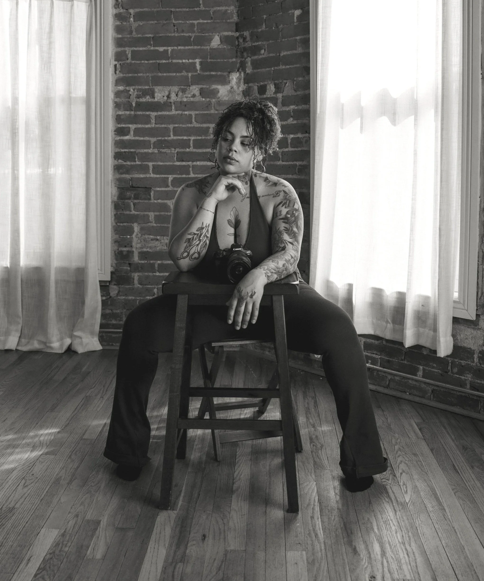 A woman with tattoos sitting on a chair in front of a brick wall, holding a camera on her lap, with her hand near her chin, in a room with wooden flooring and curtains.
