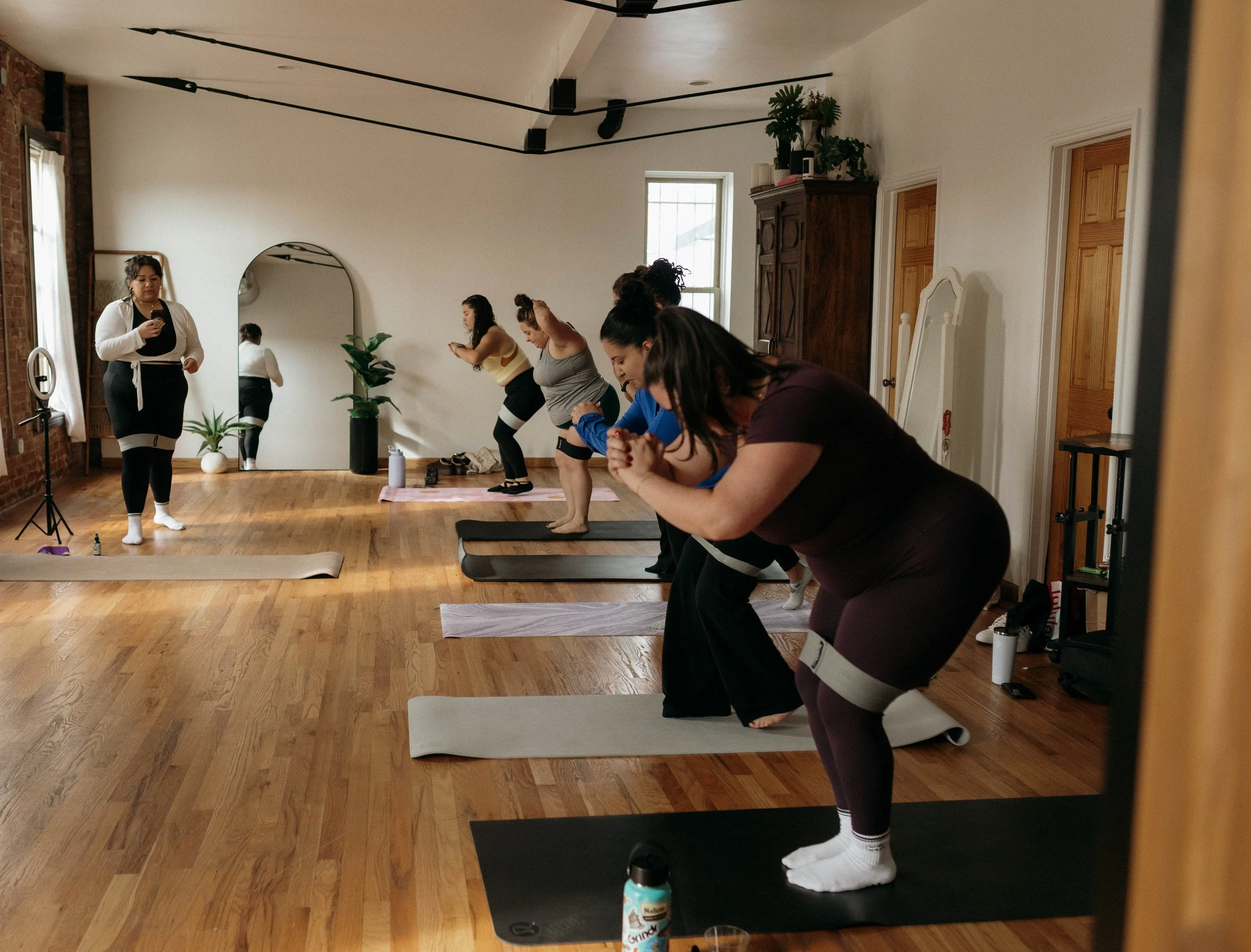 A group of women participating in a yoga class in a room with wooden floors and white walls. They are standing on yoga mats and appear to be in a forward bend pose. An instructor stands at the front near a mirror and plants, giving instructions.