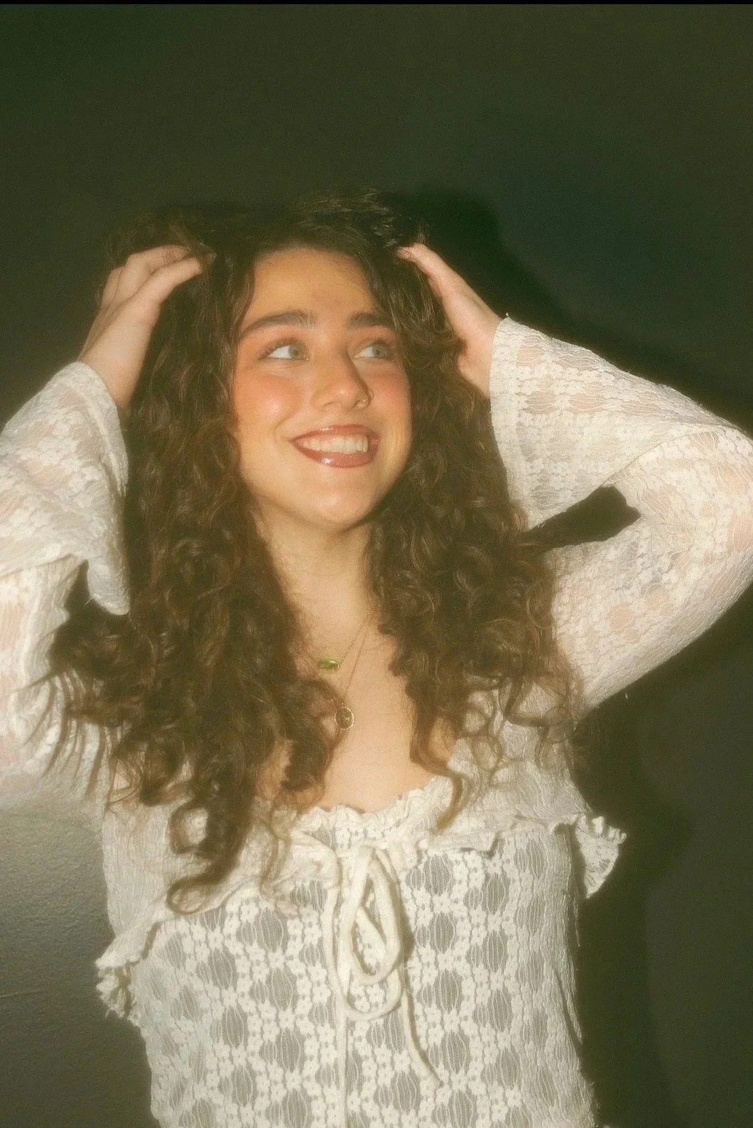 A woman with blonde hair, wearing a blue top, jeans, and beige heels, sitting on a black chair with a dark green and white wall in the background.