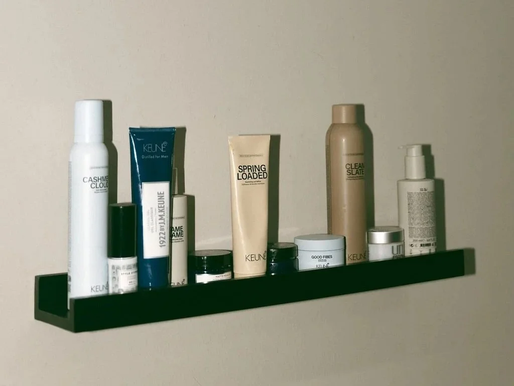 Various skincare and grooming products on a black wall-mounted shelf against a light-colored wall.