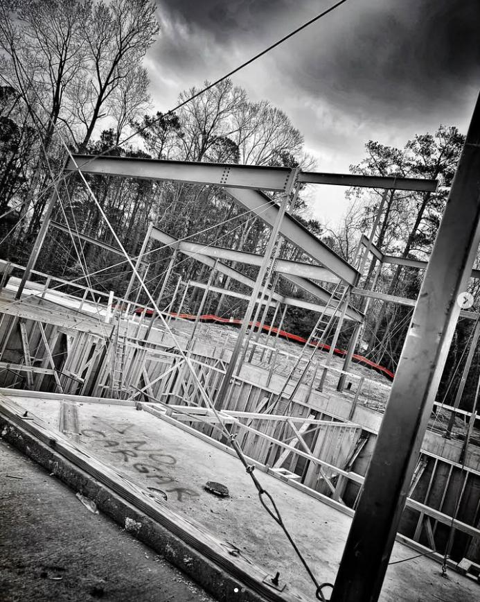 Black and white photo of a construction site with metal framework and scaffolding, set against a cloudy sky and trees in the background.