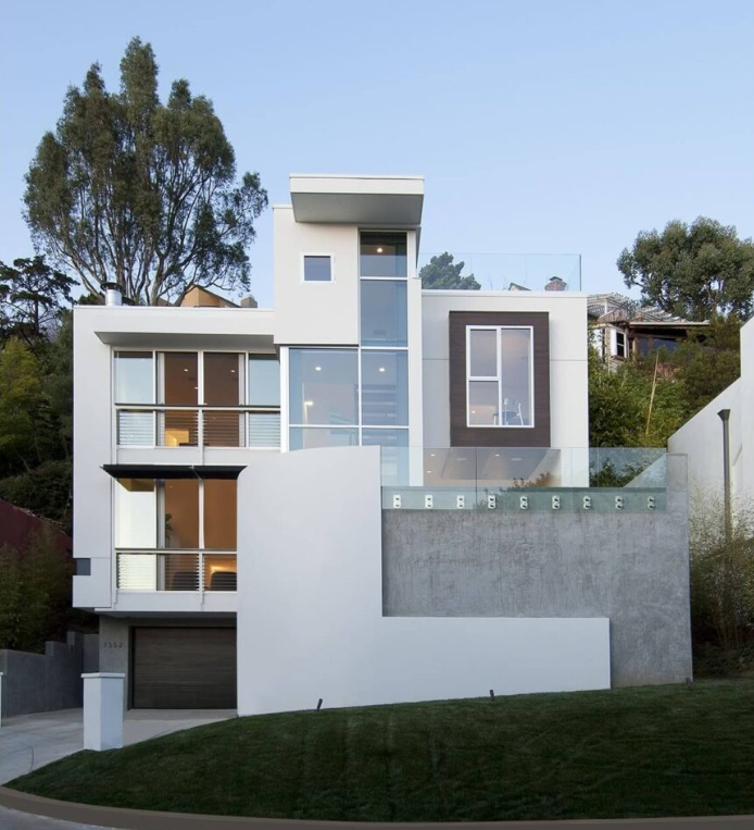 Modern multi-story house with large glass windows and a garage, situated on a hill with a grassy lawn, trees, and a blue sky in the background.