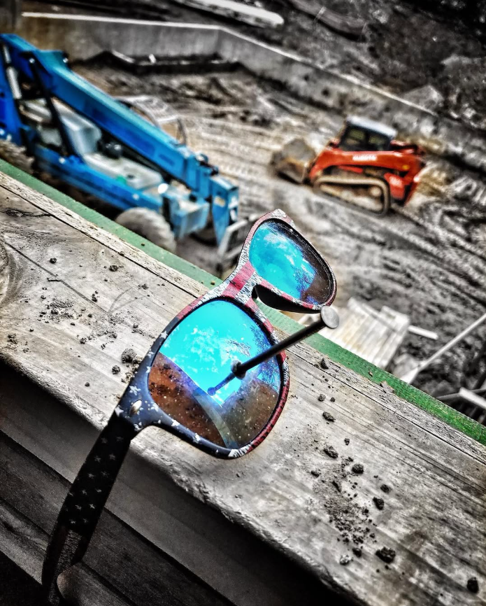 Construction site with a pair of sunglasses skewered by a nail on a wooden surface, with construction machinery in background.