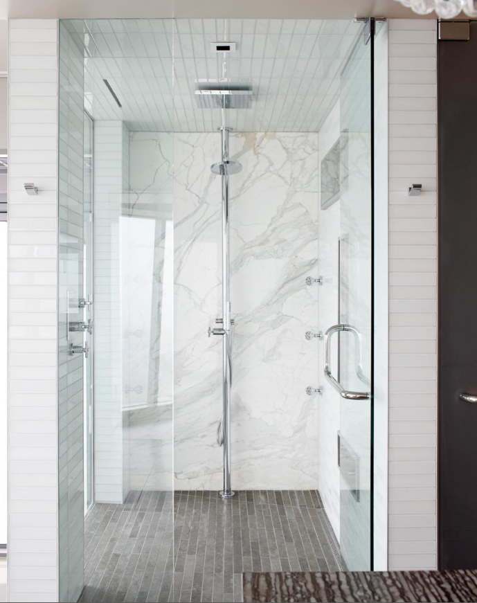 Modern glass-enclosed shower with white marble walls, gray tile floor, and a ceiling-mounted rainfall shower head.