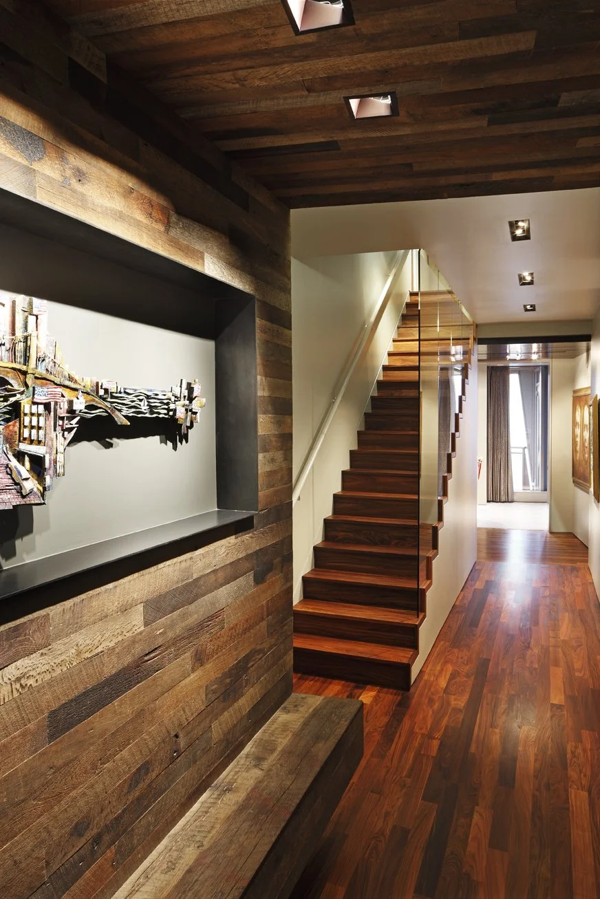 Interior view of a modern penthouse featuring reclaimed wooden walls, staircase, and flooring, with artwork on the wall and a window with curtains at the end of the hallway.