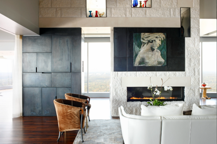 Modern living room with a white leather sofa, brown armchairs, a stone fireplace with a painting of a dog above, and large windows showing a city view.