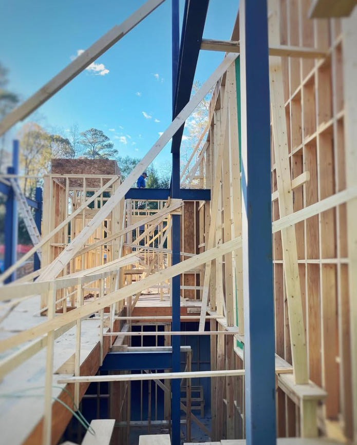 Construction site with wooden framing and steel beams for a multi-story building, exposed stairwell, and blue sky in the background.