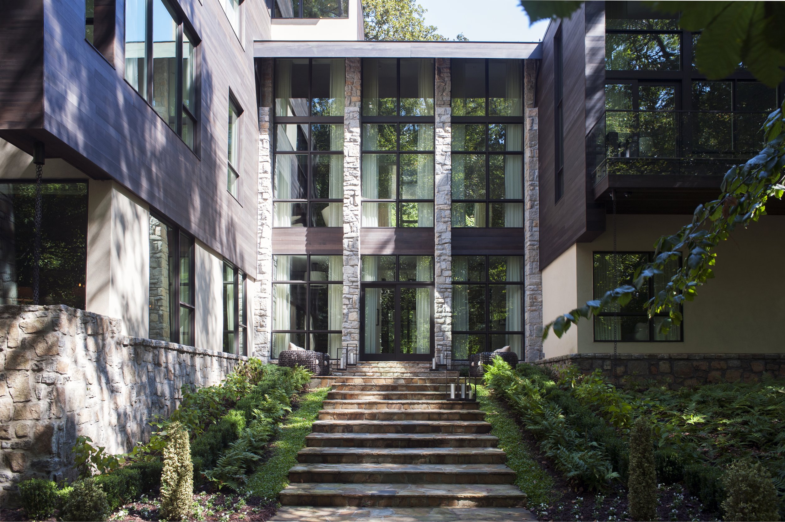 Modern multi-story building with large glass windows, stone accents, and wooden siding, surrounded by lush greenery and a stone staircase leading to the entrance.