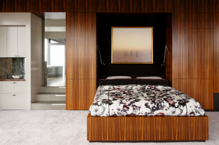 Modern penthouse bedroom with a wooden bed frame, floral bedding, framed artwork above the bed, dark wall paneling, and an adjoining bathroom with steps and a large window.
