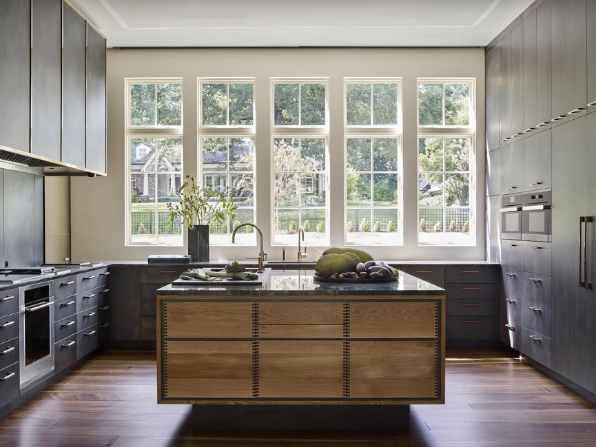 Modern kitchen with large windows, dark gray cabinets, a central island with wooden drawers, and minimal decor including a plant and vegetables on the island. Subzero-Wolf Kitchen Design Contest Global Finalist.