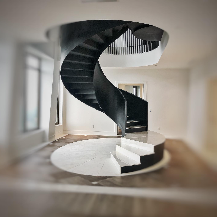 Interior view of a room with a self supporting steel spiral staircase, white walls, and a marble circle in the floor beneath the staircase.