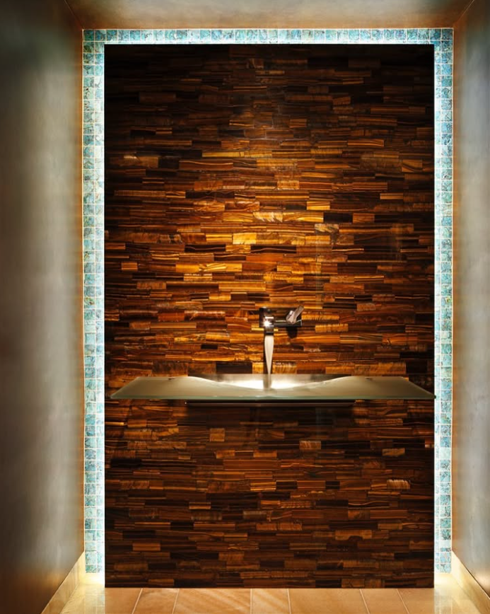 Wall-mounted glass sink with a shiny faucet against a semi precious stone slab backdrop, framed by turquoise tile on the top and sides.
