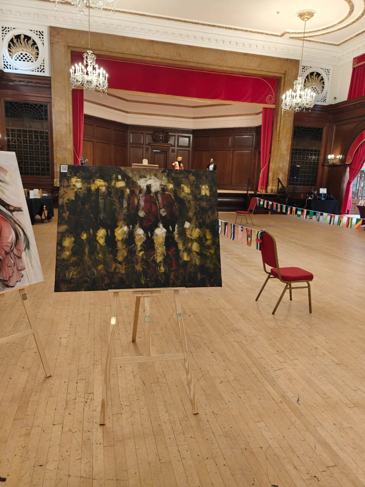 An art exhibit in a grand ballroom featuring paintings on easels, with red curtains, chandeliers, wooden paneling, and a stage with figures in traditional attire.