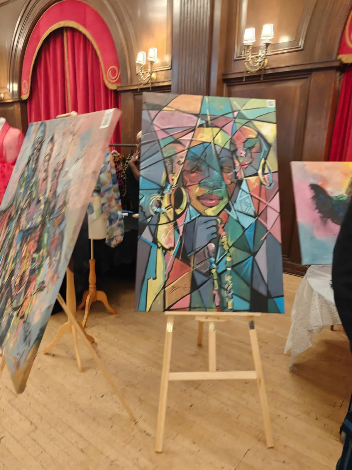 Colorful abstract painting of an African woman wearing earrings and jewelry, displayed on an easel in an elegant room with wood paneling, red curtains, and wall sconces.