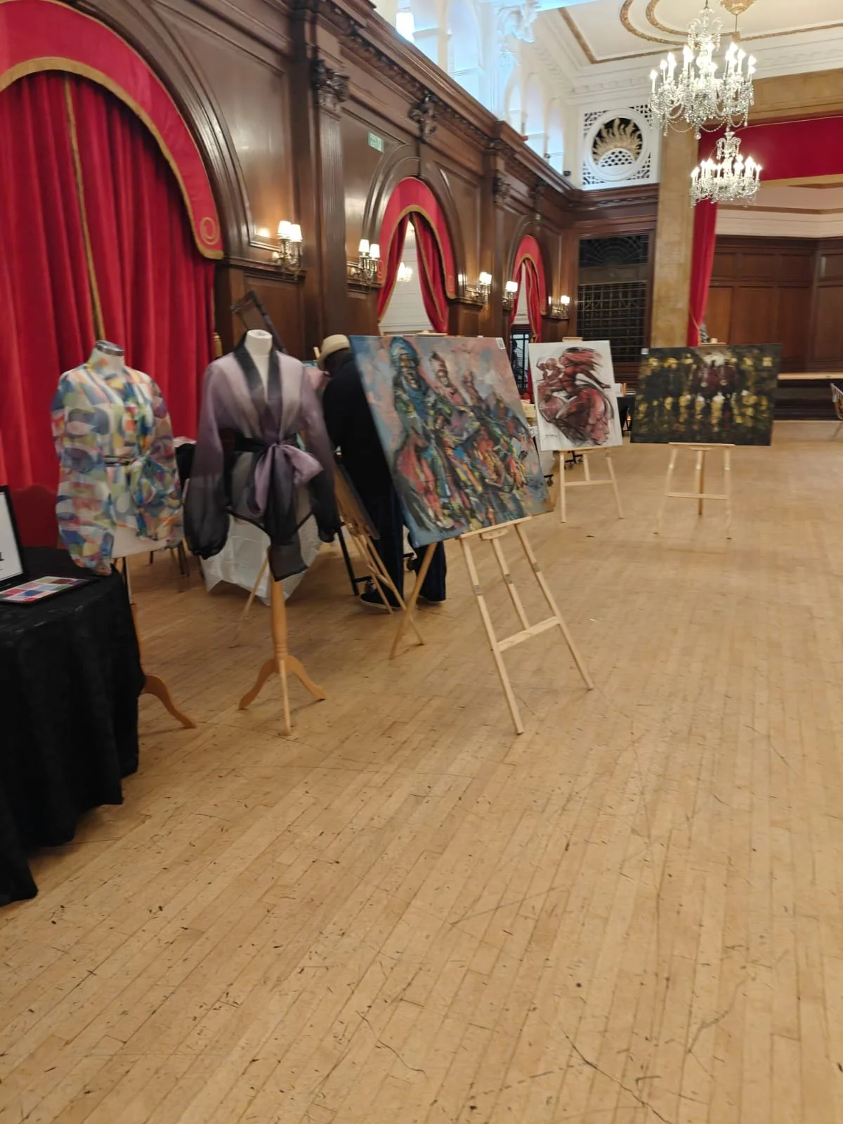 An art exhibit with paintings on easels and mannequins dressed in fashion, set in a grand hall with wood-paneled walls, red curtains, and chandeliers.
