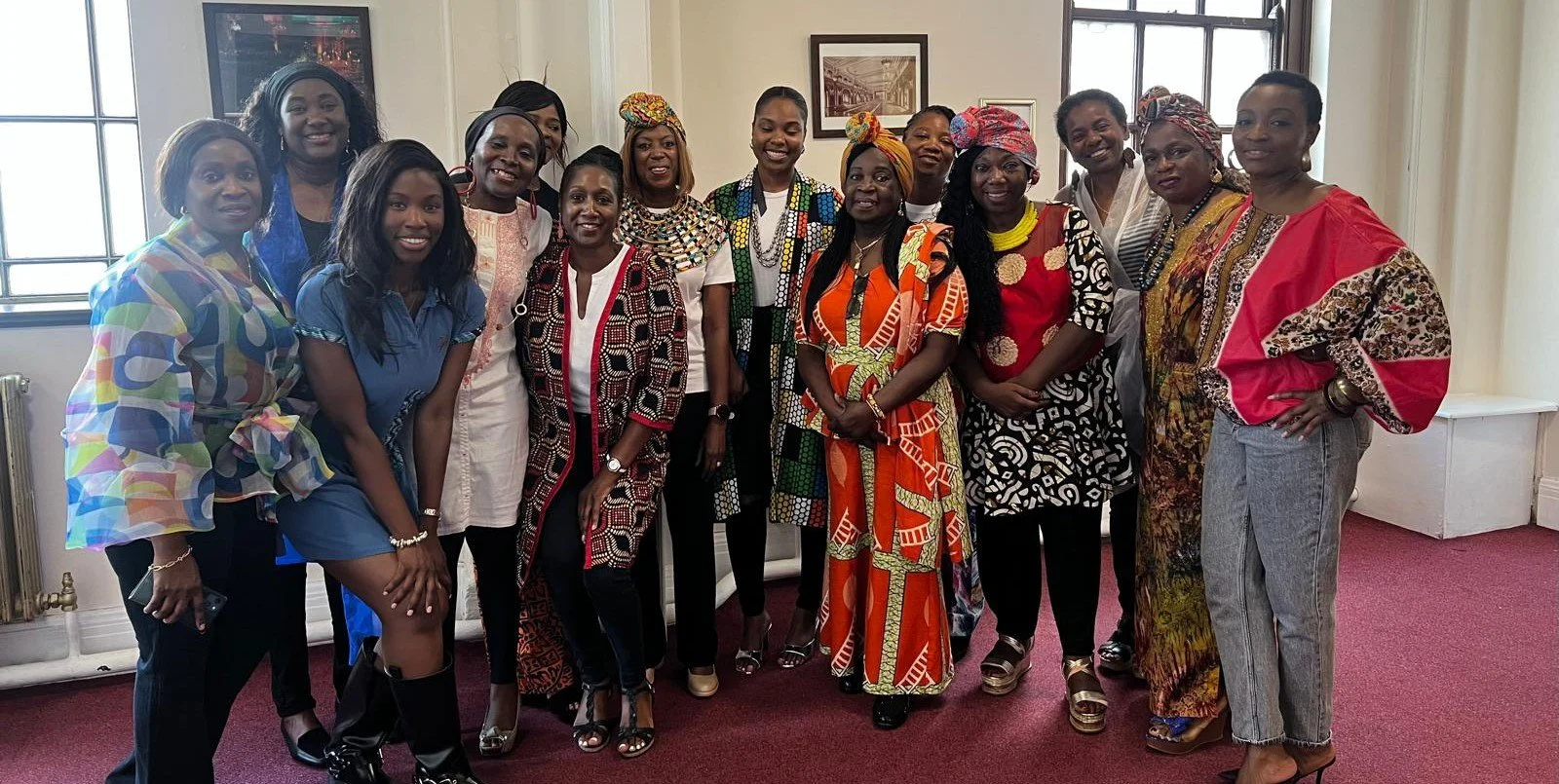 Group of women in colorful traditional and modern African clothing posing indoors with a window and framed pictures in the background.