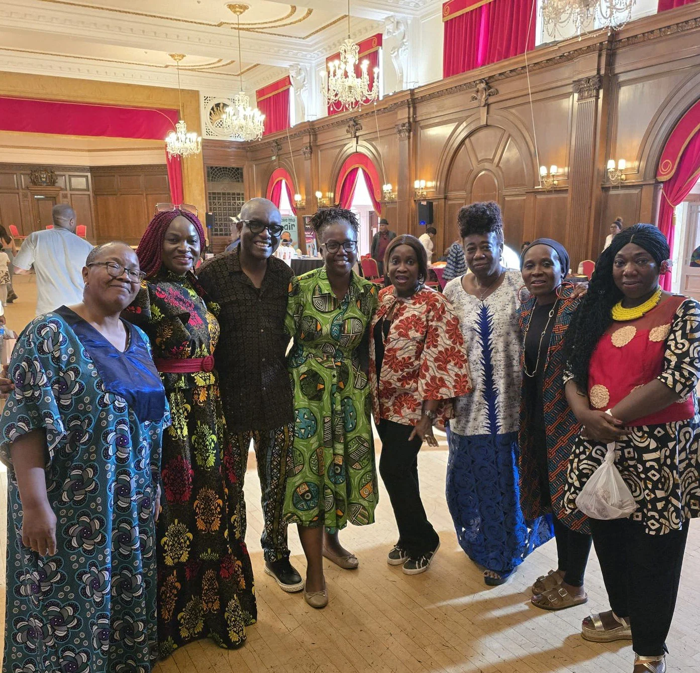 A group of nine women standing together in a grand hall with chandeliers, wooden paneled walls, and large red curtains, wearing colorful and patterned dresses.