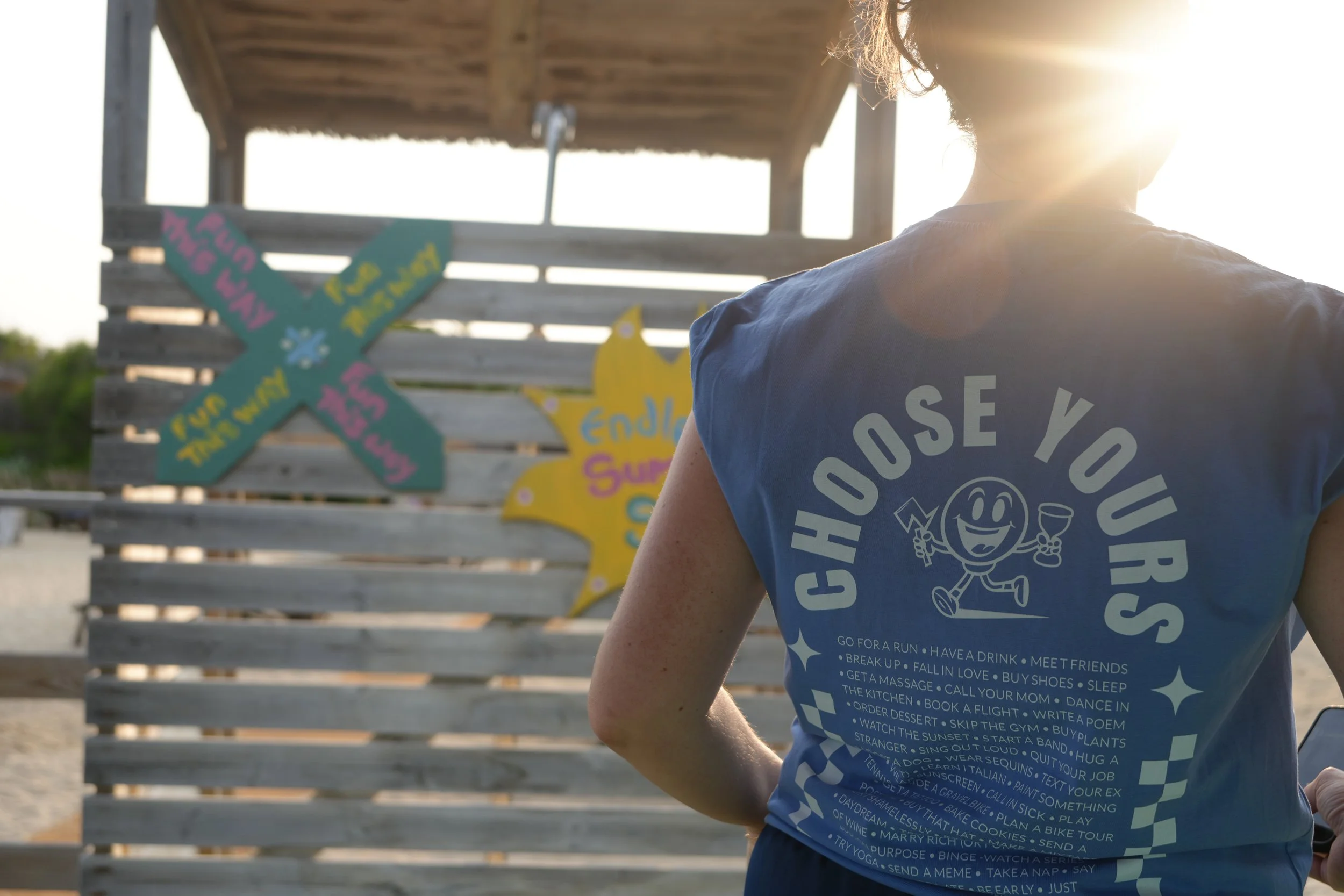 Person in blauer T-Shirt mit dem Text "Choose Yours" und einer lachenden Witzfigur auf dem Rücken, im Hintergrund bunte Schilder mit Aufschriften wie "Endless Summer" und "Make a Splash" an einer Holzpinnwand, Sonnenlicht im Hintergrund.